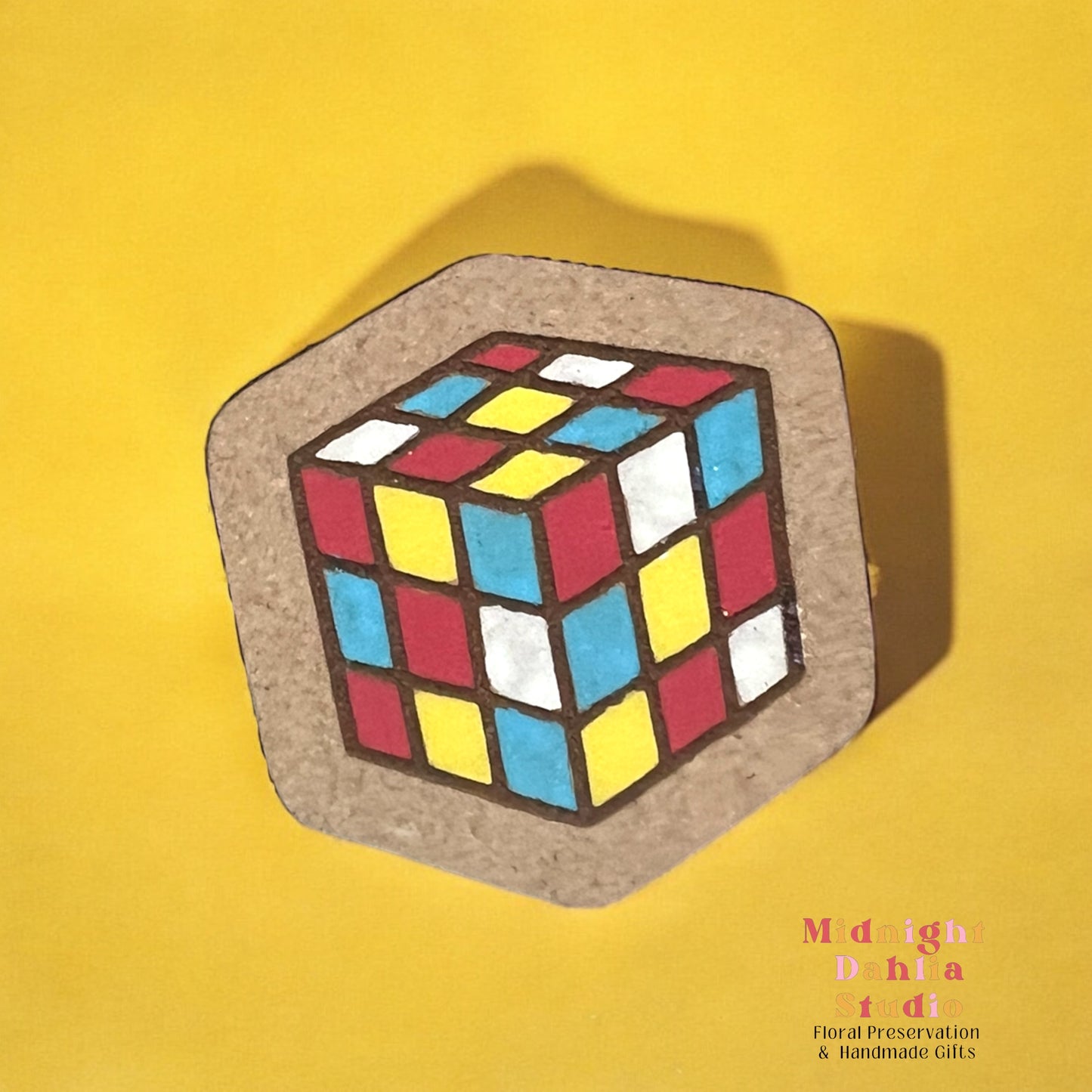 A wooden square pin with a painted Rubik’s cube with blocks of red yellow blue and white. Retro and vintage vibes.