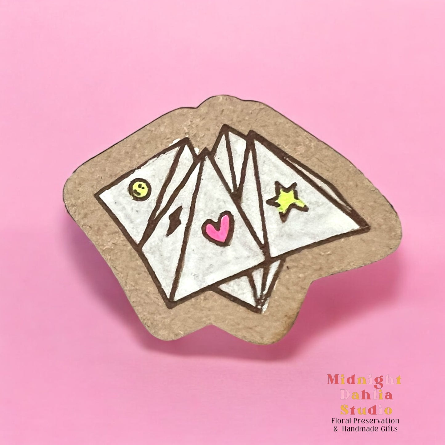 A wooden pin in the shape of a cootie catcher kid’s game in white with small bright symbols of a smile, lightning bolt, heart, and star. Retro and vintage vibes.