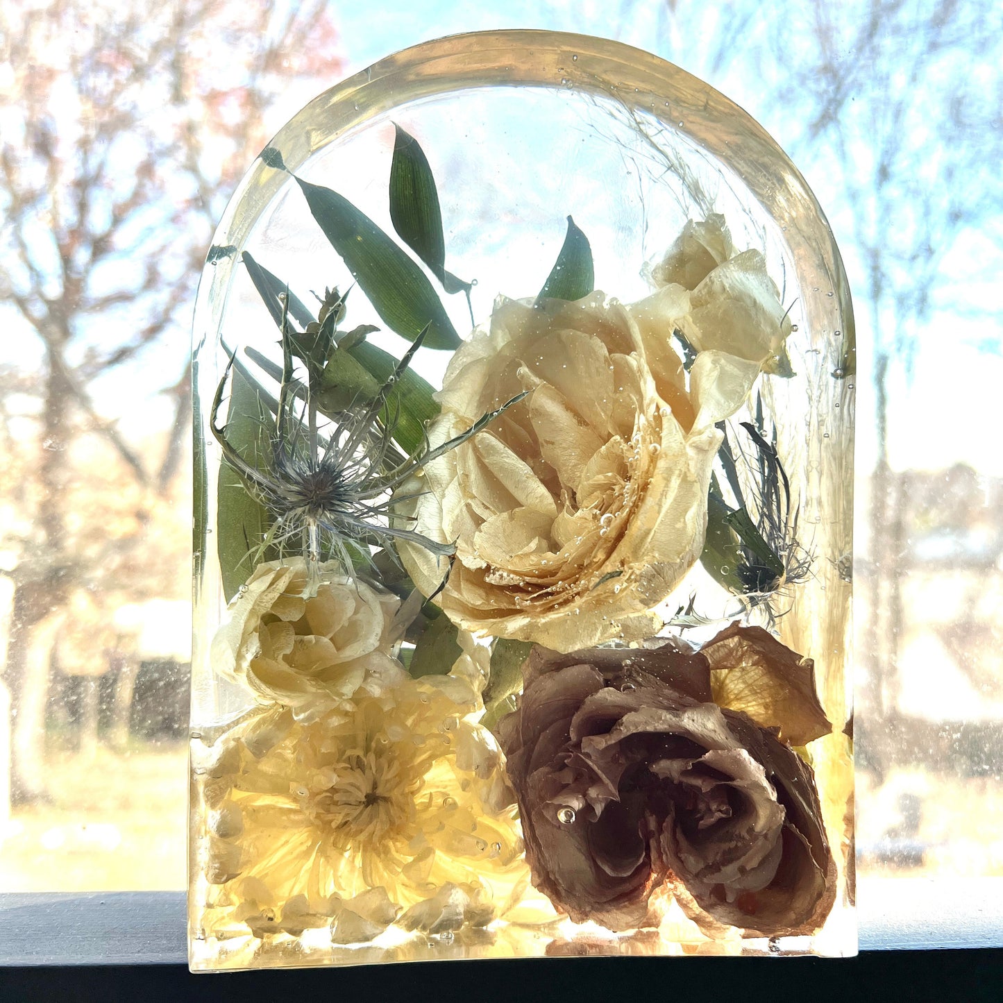 A custom crystal clear resin arch made with preserved flowers of brown taupe roses , white roses , white ranunculus , white mini spray roses , blue thistle , and green leaves . Beautiful bouquet preservation option that helps you keep your bridal flowers forever.
