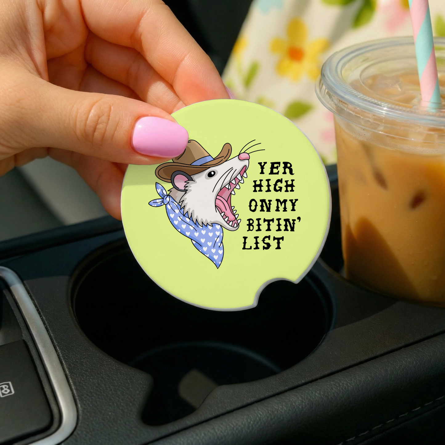 Coaster with cartoon rat design and text in a car cup holder with iced coffee.