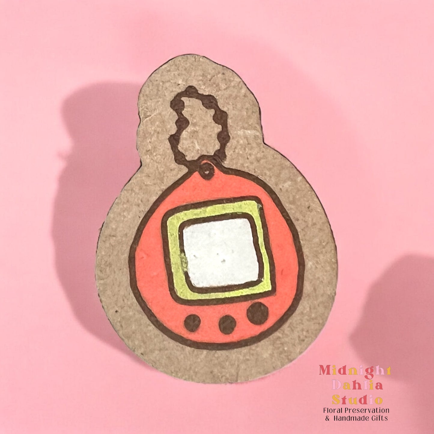 A wooden pin with a painted keychain in the style of a tamagotchi with an orange border and a small white screen surrounded by a yellow border with three buttons below the screen. Retro and vintage vibes.