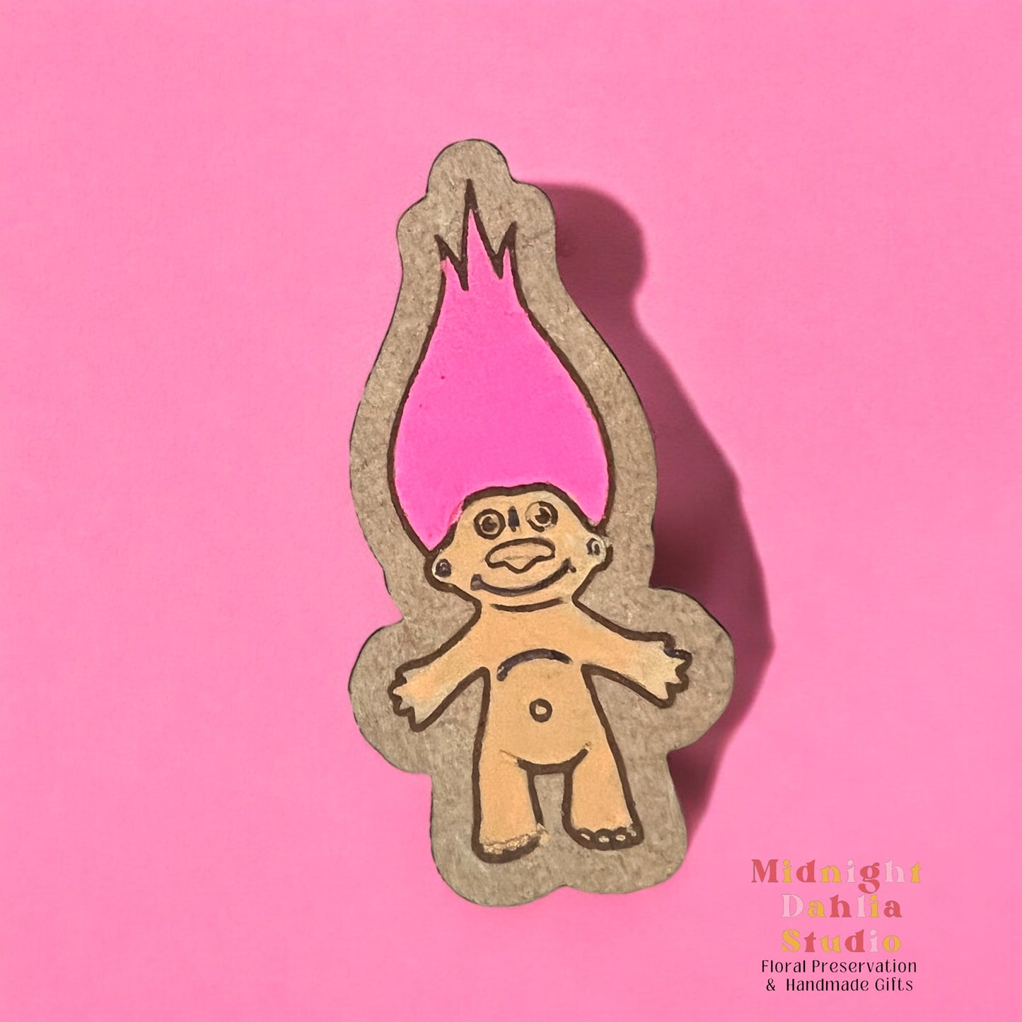 A wooden pin with a hand painted troll doll with peach skin and bright pink hair. Retro and vintage vibes.