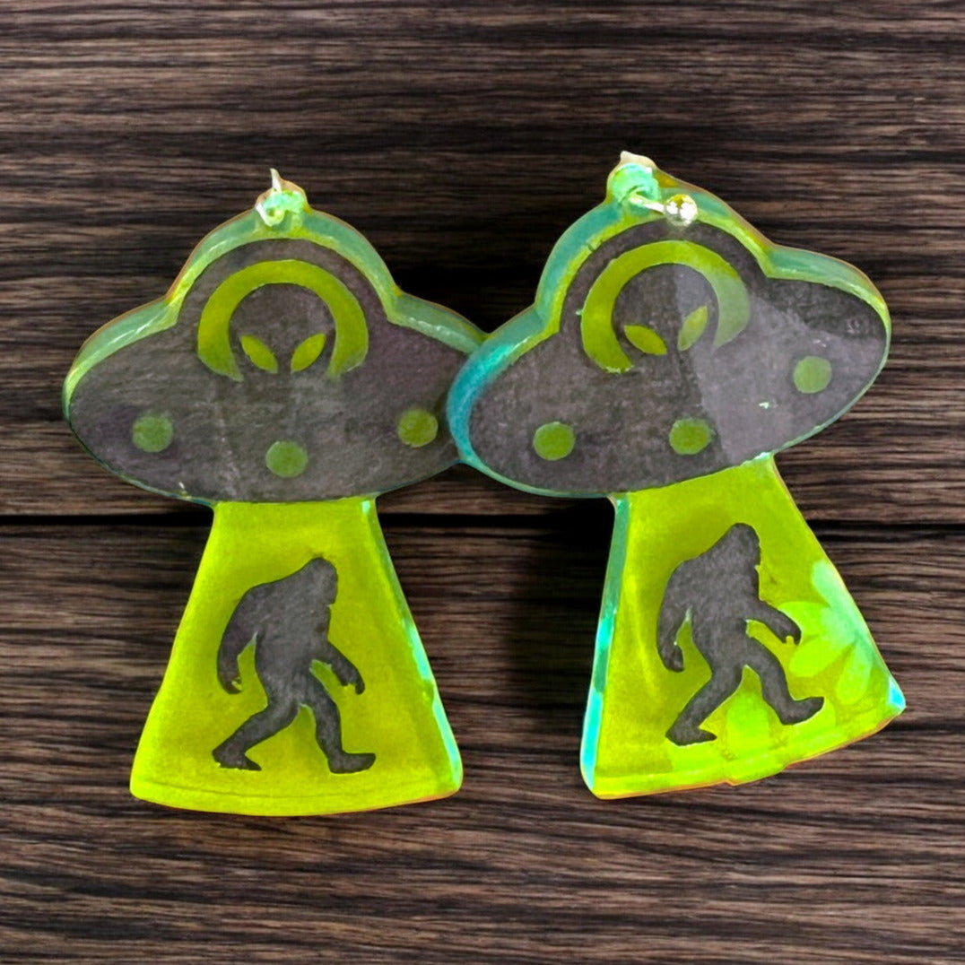 A pair of dangle earrings that depict a ufo with an alien driver abducting a bigfoot. the earrings are made of neon green acrylic and are a perfect gift for a woman that loves cryptids and silliness.