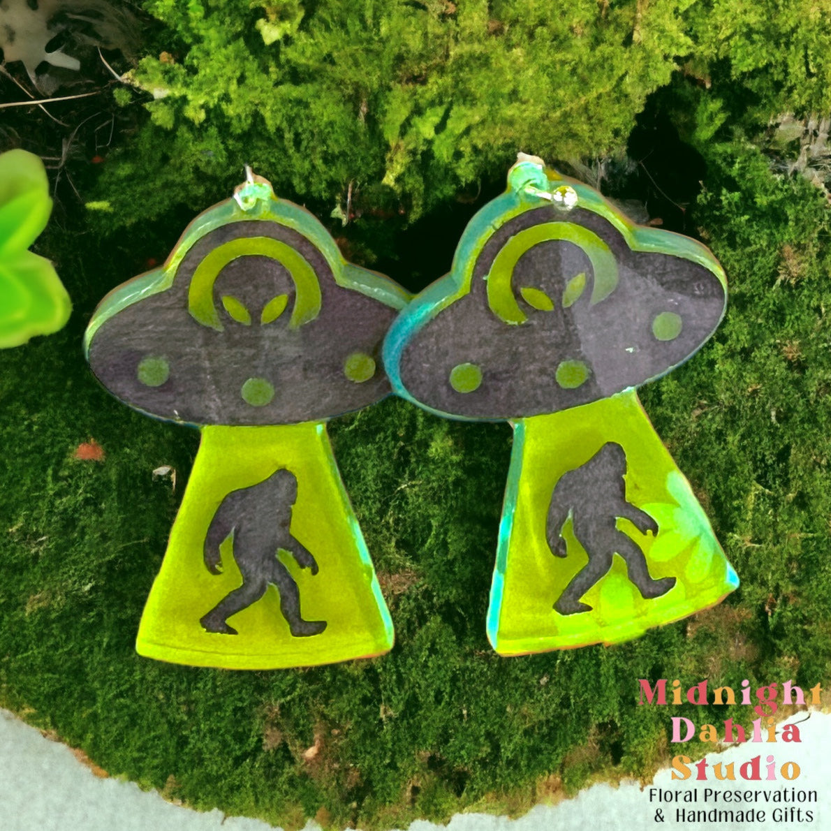 A pair of dangle earrings that depict a ufo with an alien driver abducting a bigfoot. the earrings are made of neon green acrylic and are a perfect gift for a woman that loves cryptids and silliness.
