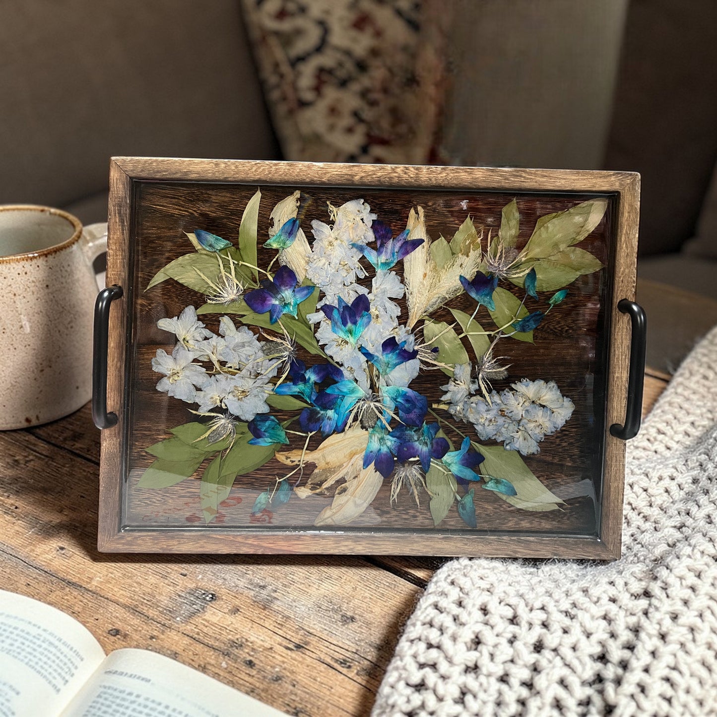 Decorative tray with pressed flowers on a wooden surface next to a mug and book.