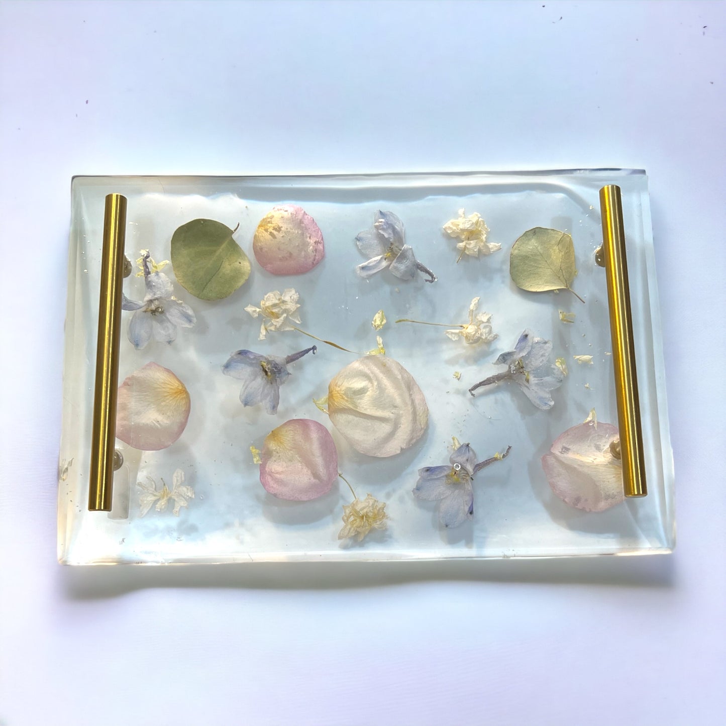 A custom serving tray with pressed flowers including pink rose petals , blue delphinium , and white larkspur . It has silver handles and is made of crystal clear resin.
