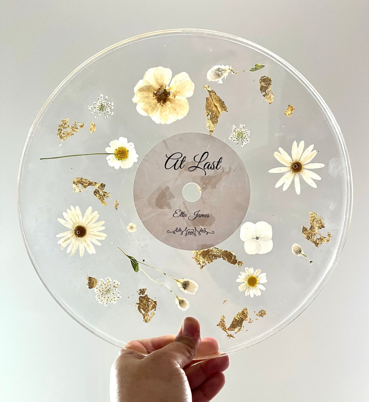 A custom decorative resin record made with white daisies , white roses , white hydrangea , and green leaves , and gold flake. It has a label to make it look like an album.