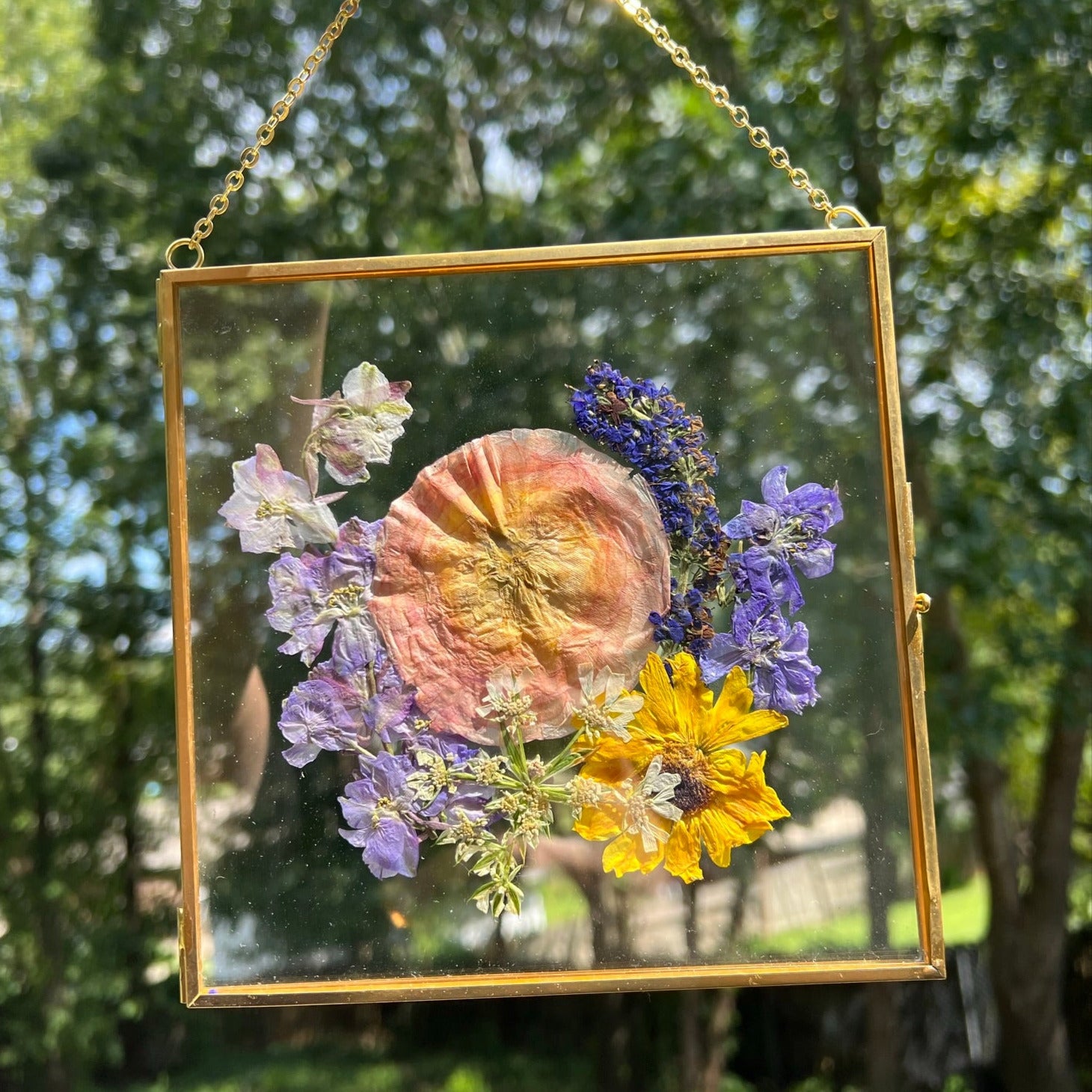 A custom pressed flower floating frame with preserved pink ranunculus , purple veronica , blue delphinium, and yellow daisies .   A beautiful way to preserve a bridal bouquet.