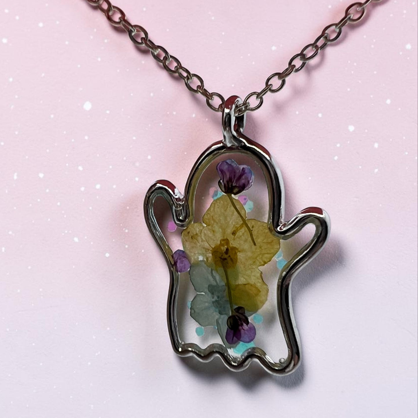 A silver outline pendant necklace shaped like a ghost, containing dried yellow and purple flowers suspended in a clear resin, shown on a pink glittery background.