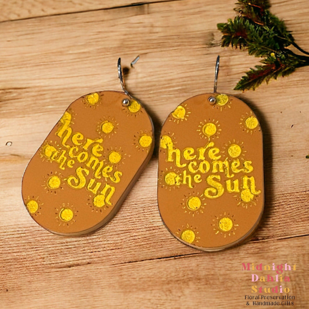 oval acrylic earrings made with orange acrylic with yellow suns and the words here comes the sun. the jewelry is on silver-plated hardware. a perfect retro gift for a woman.