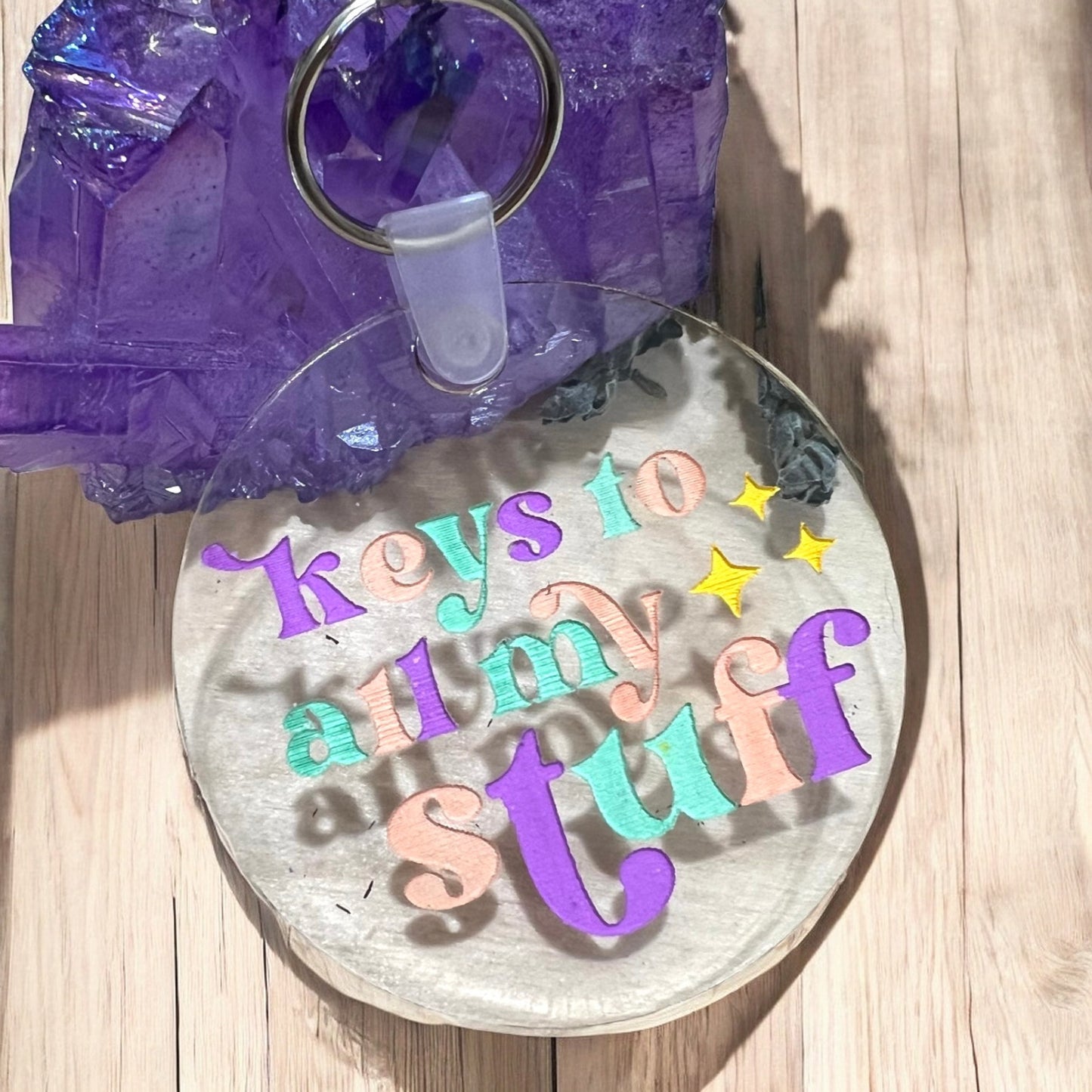 A round clear acrylic keychain with keys to all my stuff etched in purple, peach orange, and teal blue green with yellow stars to the right.