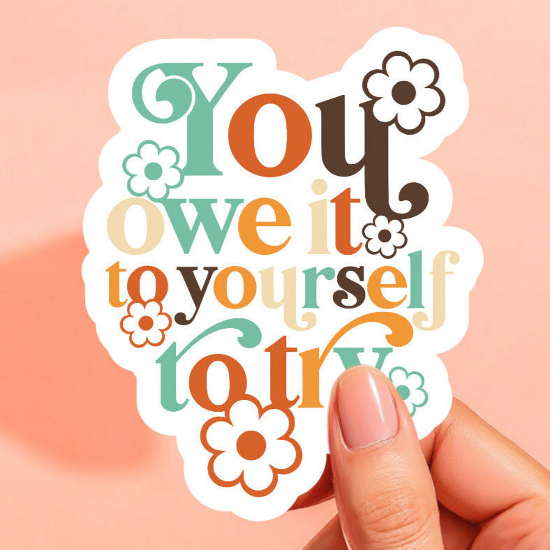 a white sticker that reads you owe it to yourself to try in colorful retro lettering surrounded by daisies in matching colors