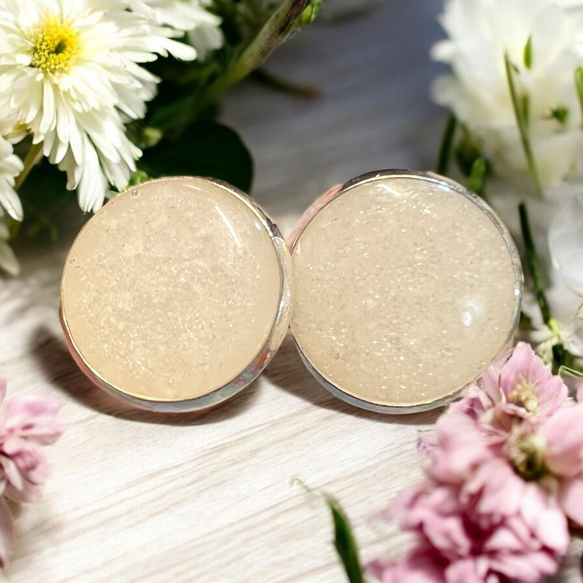 Two round cosmetic products with a textured surface, surrounded by flowers on a light wooden surface.
