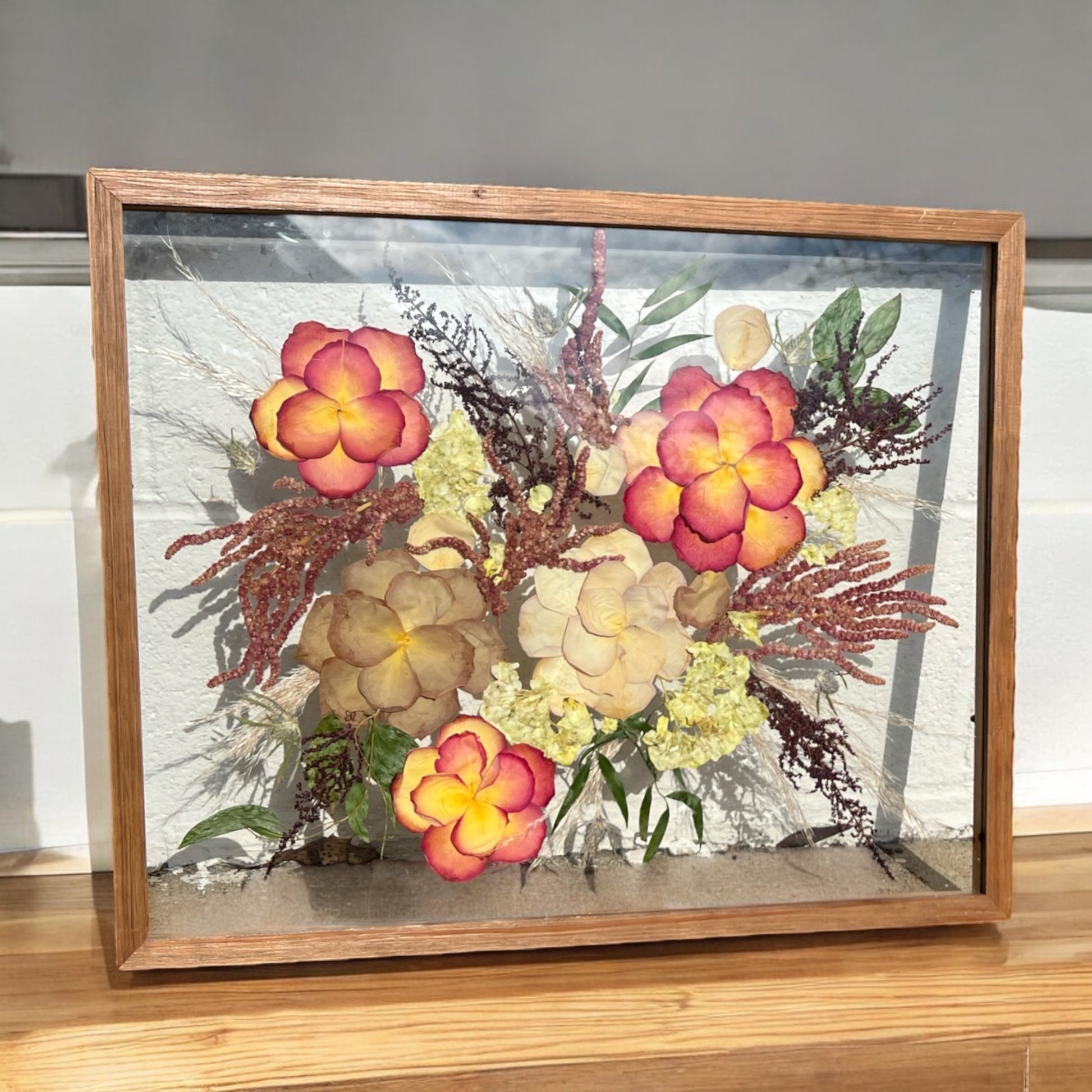 A custom pressed flower floating frame with preserved orange and yellow roses , mauve taupe roses , red amaranthus , burgundy red astilbe , and green eucalyptus .   A beautiful way to preserve a bridal bouquet.