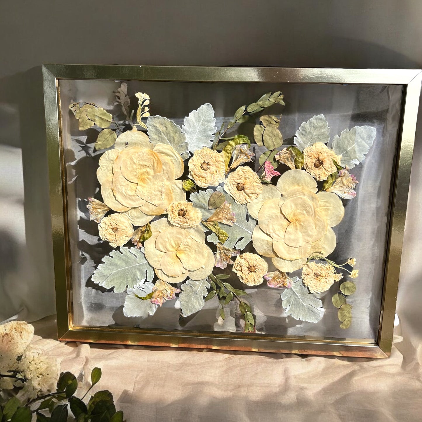 A custom pressed flower floating frame with preserved white roses , pink flowers , and dusty miller leaves . A beautiful way to preserve a bridal bouquet.