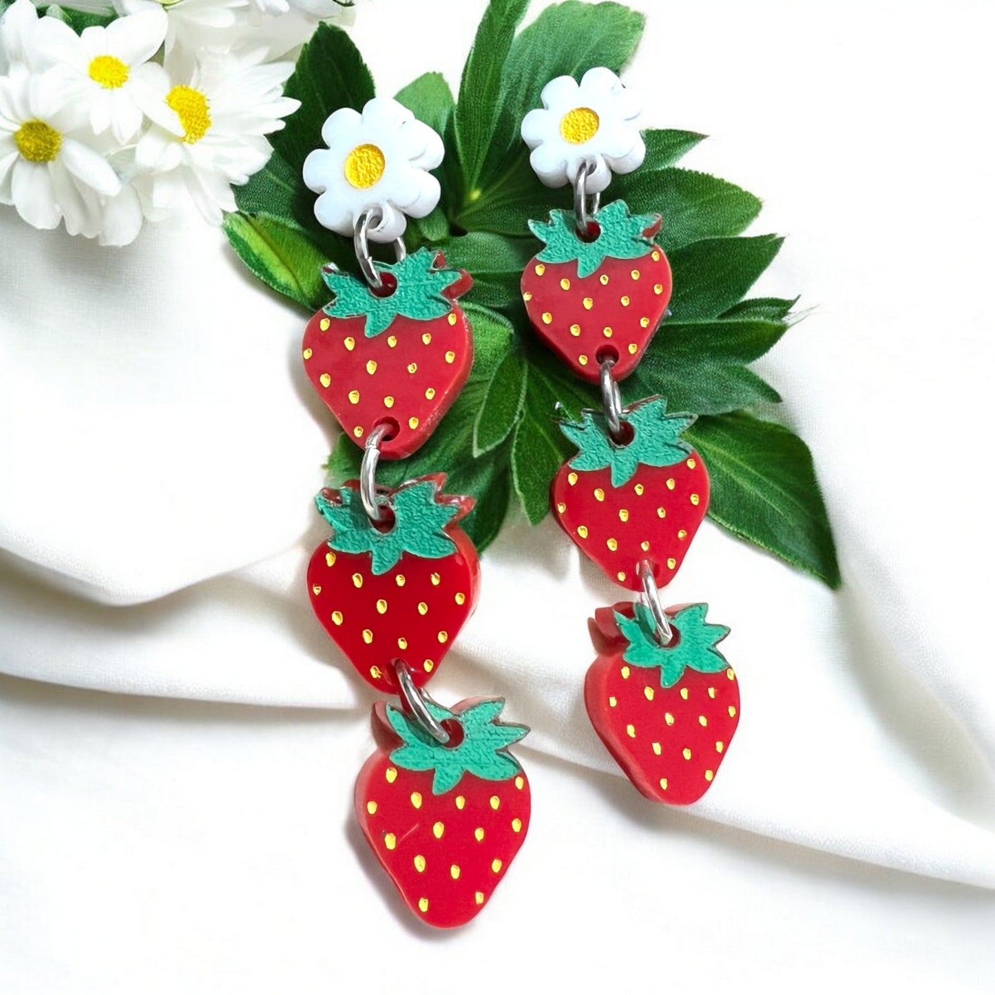 A pair of earrings made of red acrylic in the shape of 3 three strawberries hanging from a white daisy with a yellow center. A perfect summer gift for a woman or teen.