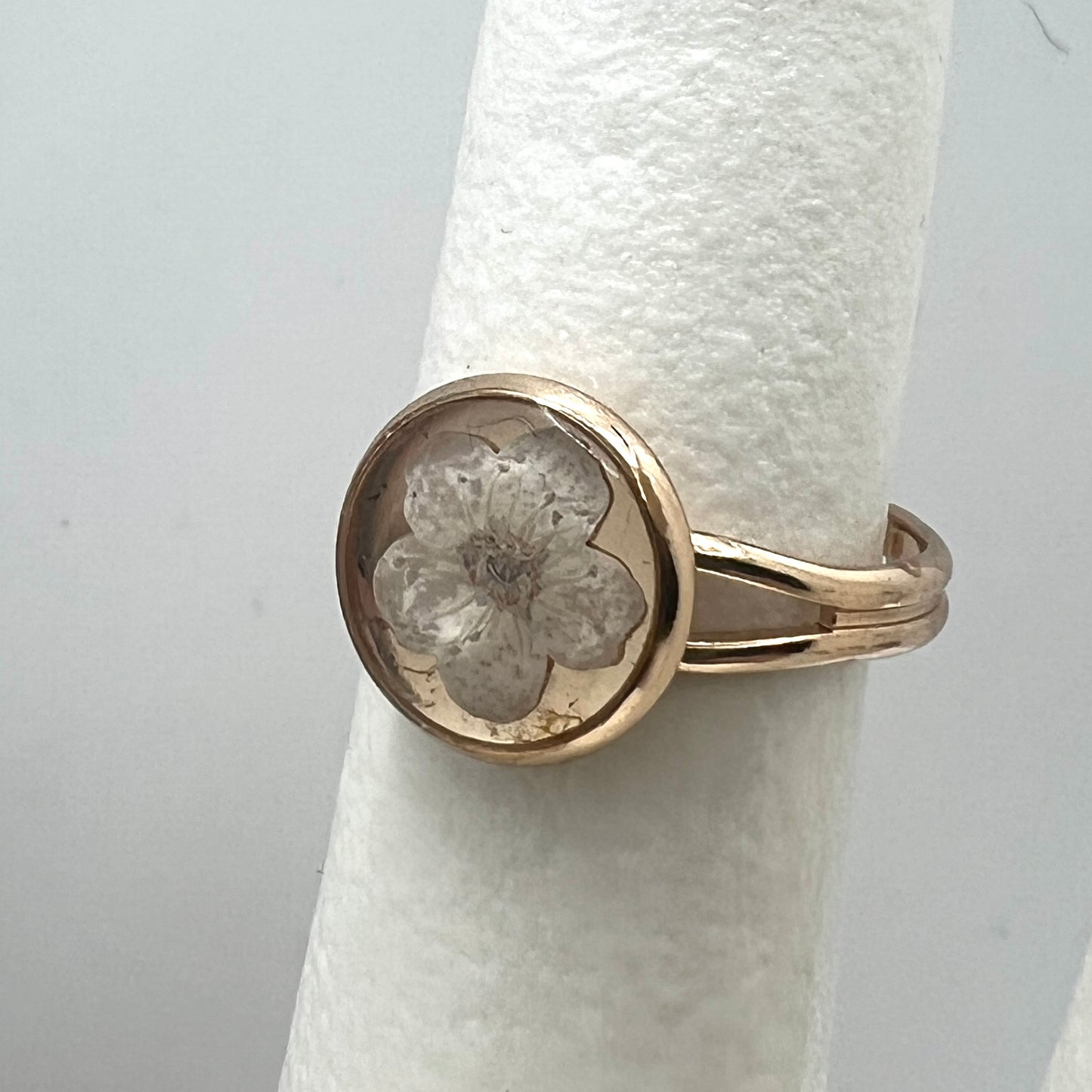 Gold ring with a floral design on a white cylindrical stand against a light gray background