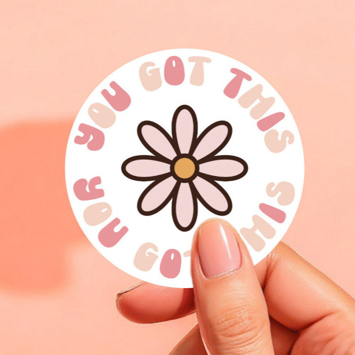 A round white sticker that reads you got this surrounding a pink daisy