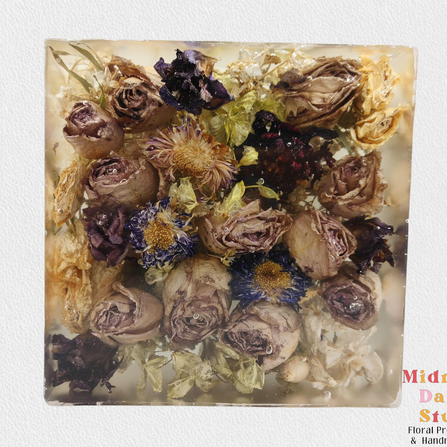 A crystal clear resin block with a lavender roses , purple strawflower, and other flowers . A beautiful way to preserve a bridal bouquet.