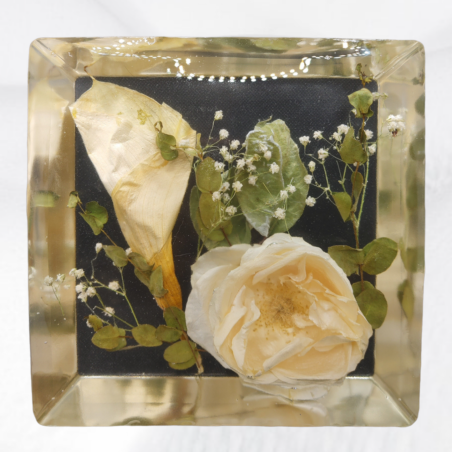 A crystal clear resin block with a white rose , white calla lily , babys breath , and leaves . A beautiful way to preserve a bridal bouquet.