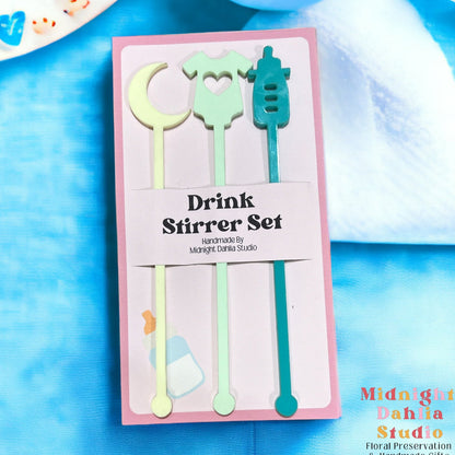 a set of three drink stirrers in the shape of a moon, a onesie, and a baby bottle in pale light yellow, light pale green, and teal turquoise blue green.