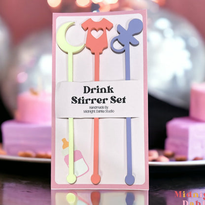 set of three drink stirrers with the top in the shape of a moon, a onesie, and a pacifier in pale light yellow, coral pink, and lavender purple.