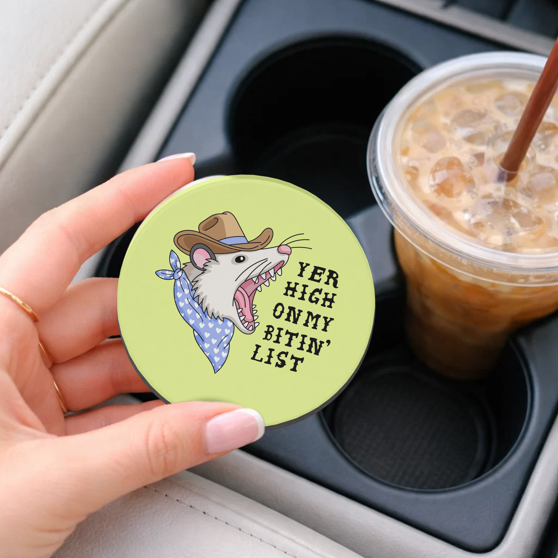 Coaster with cartoon rat design and text held by a hand over a cup holder with iced coffee.