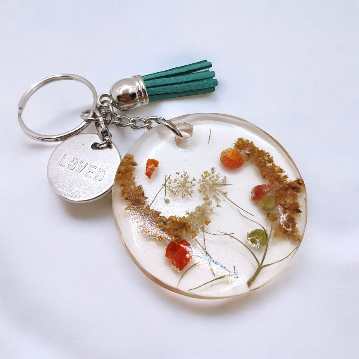 a round resin keychain with fall flowers embedded in it on silver hardware with a teal tassel against a white background