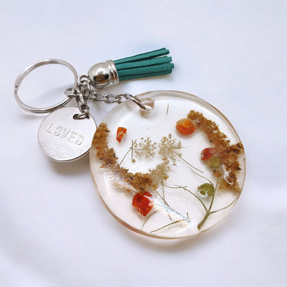 a round resin keychain with fall flowers embedded in it on silver hardware with a teal tassel against a white background