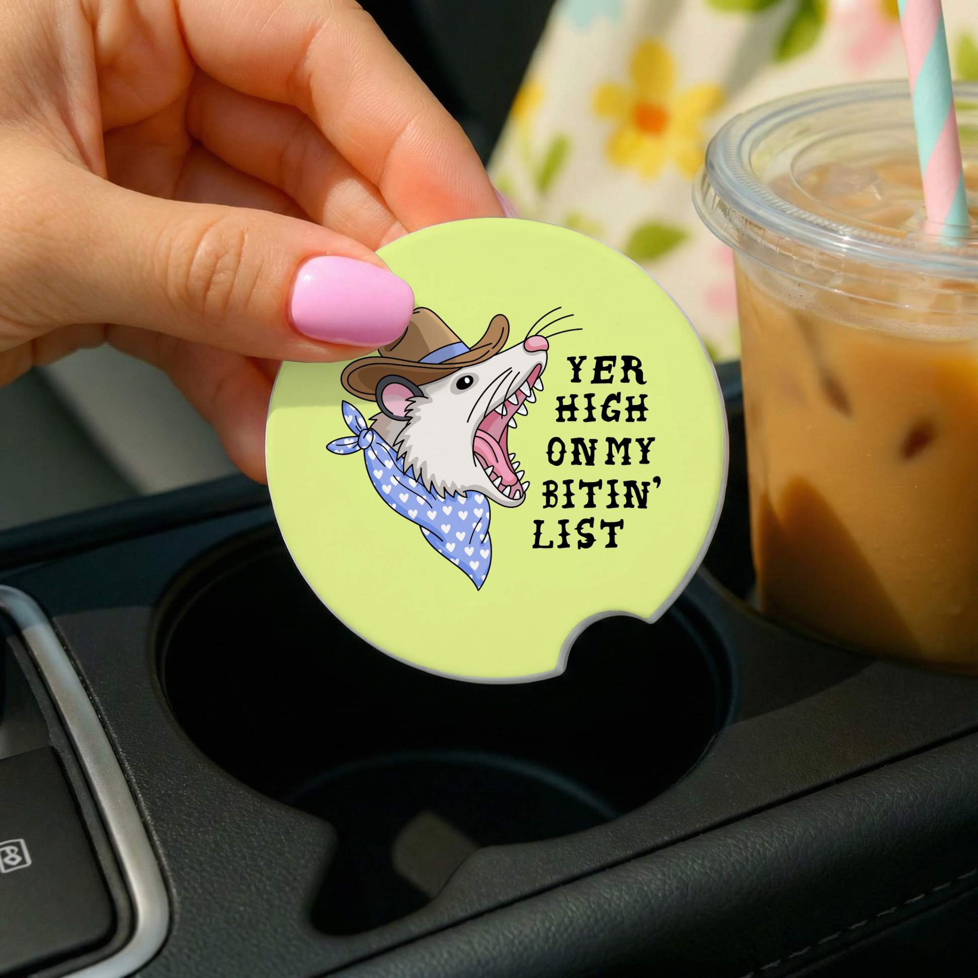 Coaster with cartoon rat design and text in a car cup holder with iced coffee.