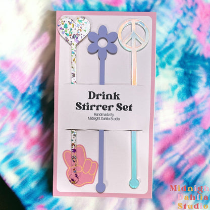 a set of three drink stirrers with the top in the shapes of a heart, daisy, and peace sign in rainbow glitter, lavender purple, and iridescent.