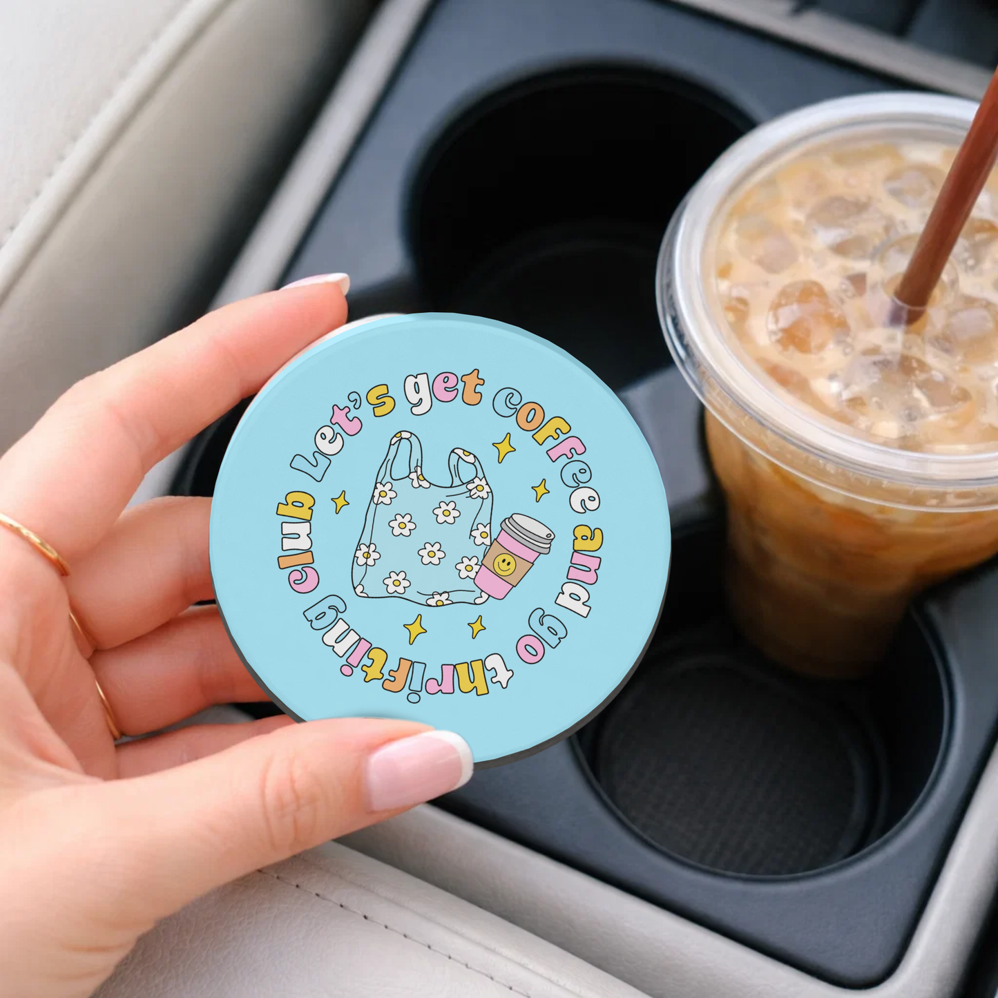 Hand holding a colorful coaster with text and graphics, next to a cup of iced coffee in a car.