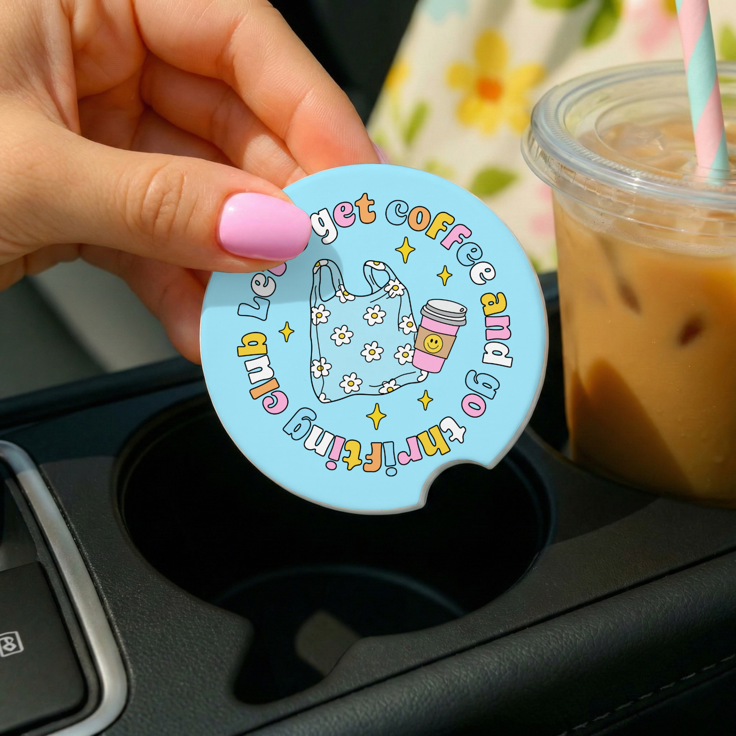Hand holding a blue circular coaster with text and graphics, placed on a car cup holder with iced coffee in the background.