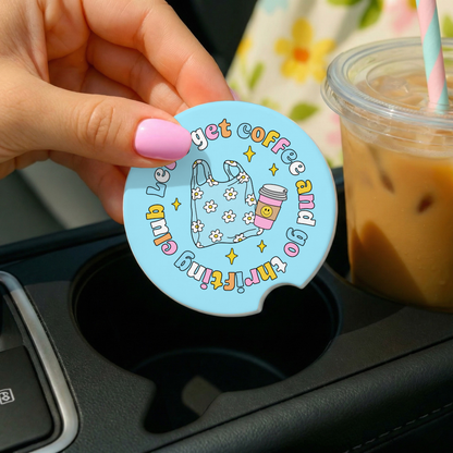 Hand holding a blue circular coaster with text and graphics, placed on a car cup holder with iced coffee in the background.