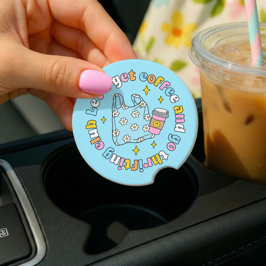 Hand holding a blue circular coaster with text and graphics, placed on a car cup holder with iced coffee in the background.
