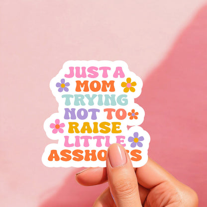 Colorful sticker with the text 'JUST A MOM TRYING NOT TO RAISE LITTLE ASUSLIES' arranged in a playful layout with floral accents.