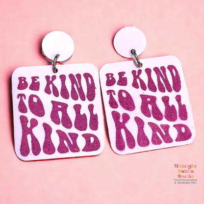 a set of two pink square acrylic earrings that read 'be kind to all kind' in a darker pink retro font