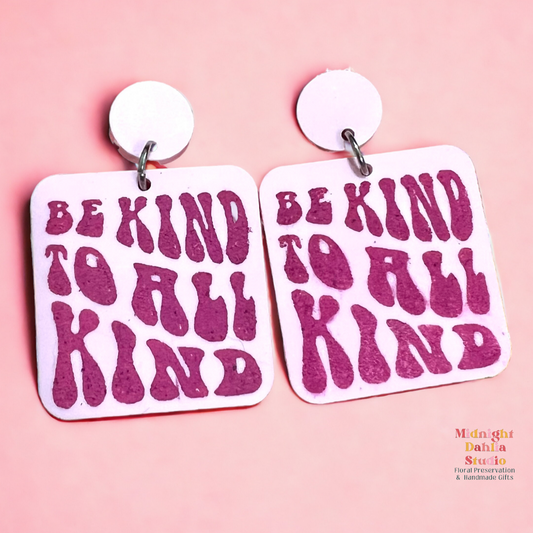 a set of two pink square acrylic earrings that read 'be kind to all kind' in a darker pink retro font