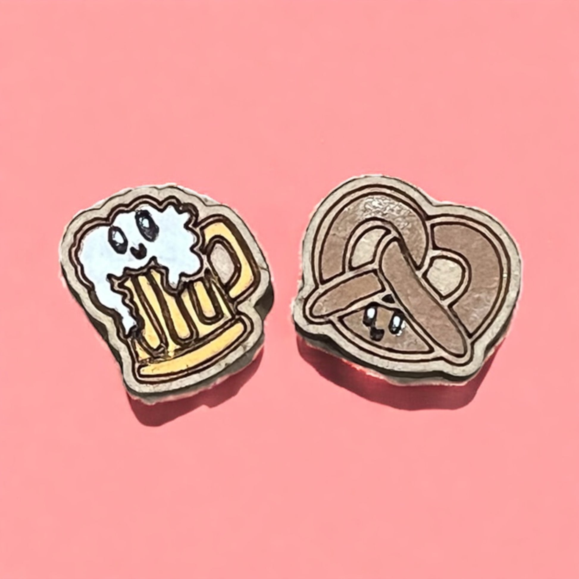 Two stud earrings with cartoon designs of a beer mug and a pretzel on a pink background.