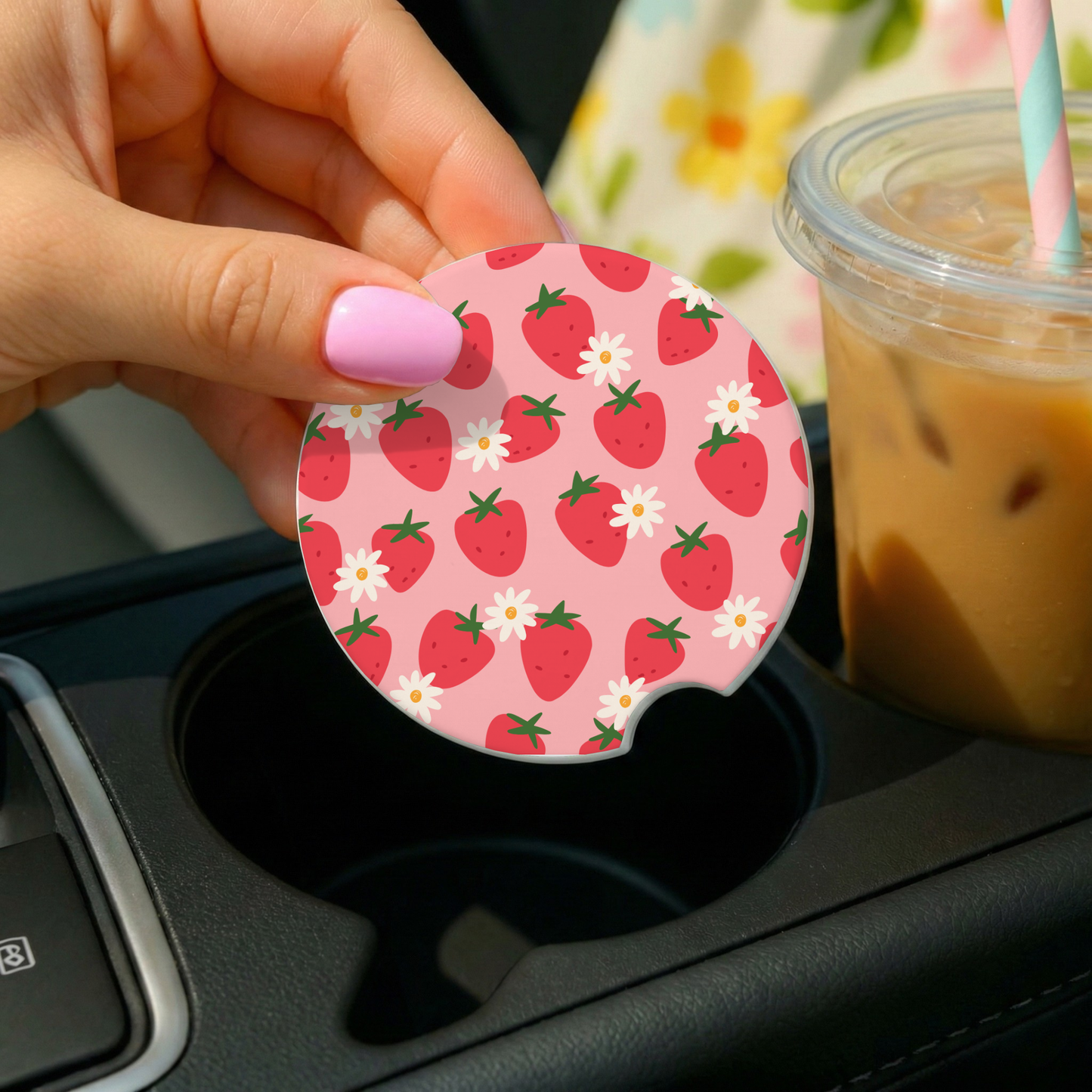 Hand holding a strawberry-patterned car coaster next to a cup of iced coffee.