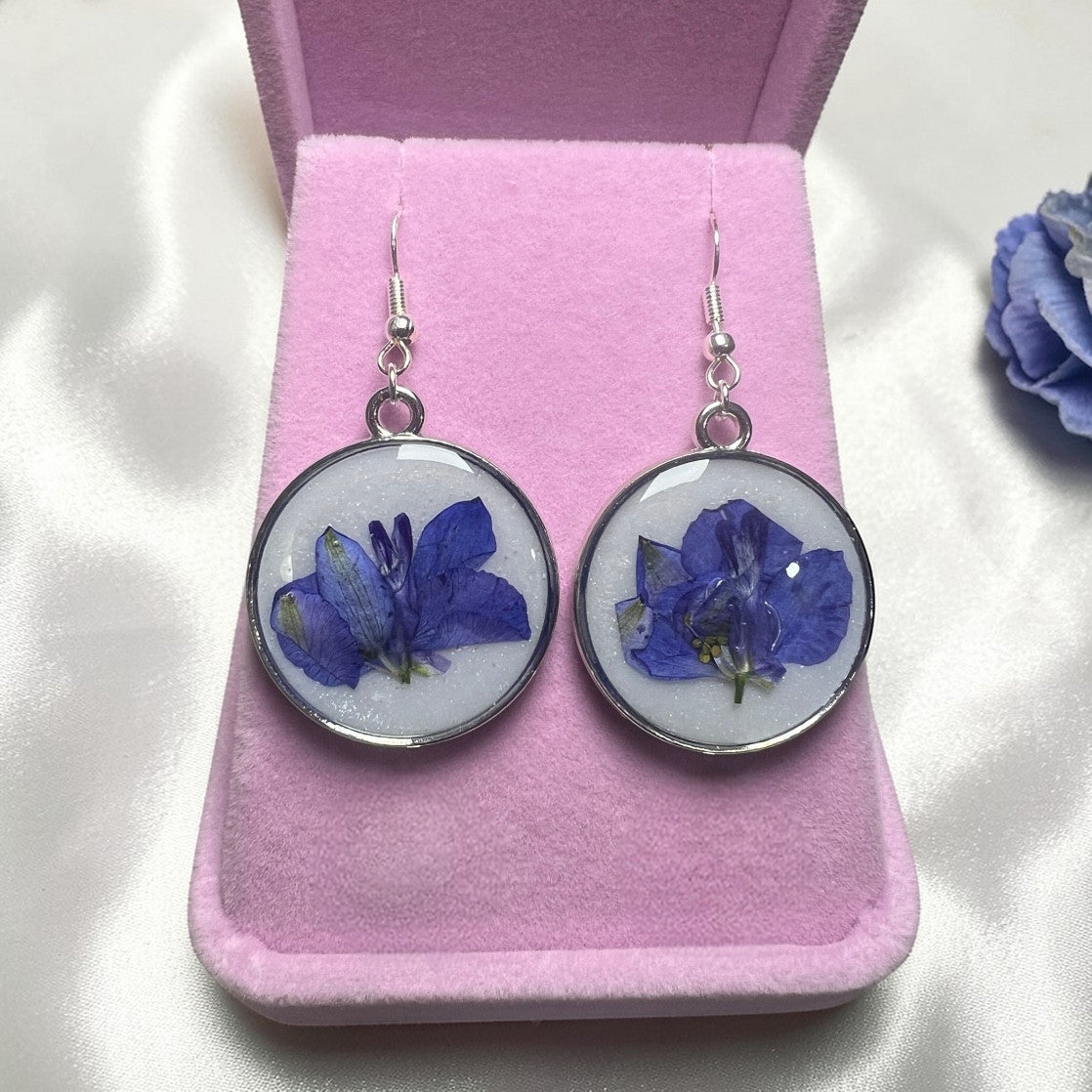 Pair of earrings with blue flower designs on a pink velvet stand.
