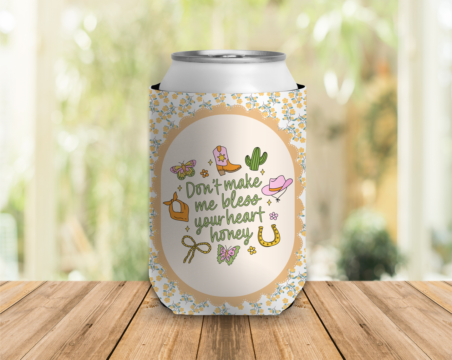 Can cooler with cowboy boot, cactus, and horseshoe design on a wooden surface with blurred greenery background