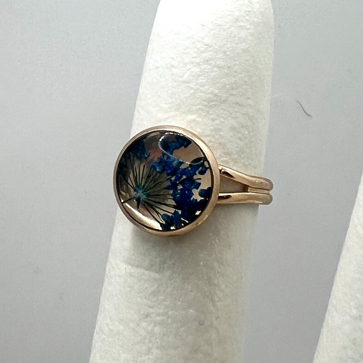Rose gold ring with a circular design featuring blue flowers on a white stand against a gray background