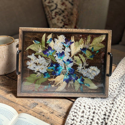 Decorative tray with pressed flowers on a wooden surface next to a mug and book.