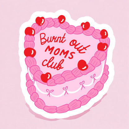 A sticker with a colorful cake design and the text 'Burnt Out Moms Club' on a pink background, held between fingers.