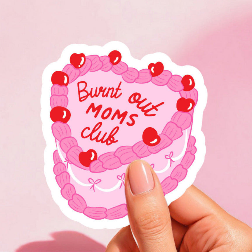 A sticker with a colorful cake design and the text 'Burnt Out Moms Club' on a pink background, held between fingers.