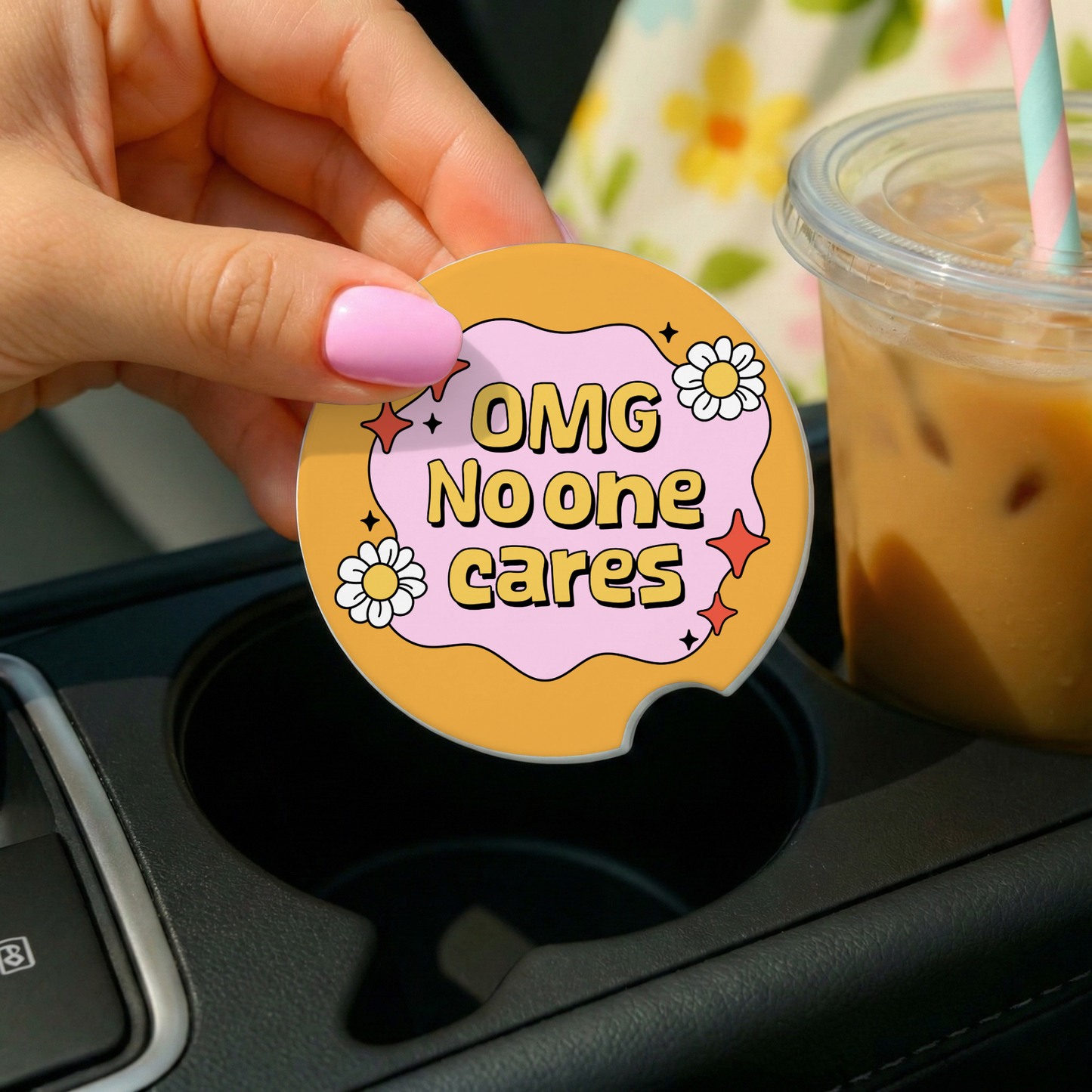 Round sticker with 'OMG No one cares' text on a car cup holder with iced coffee in the background.