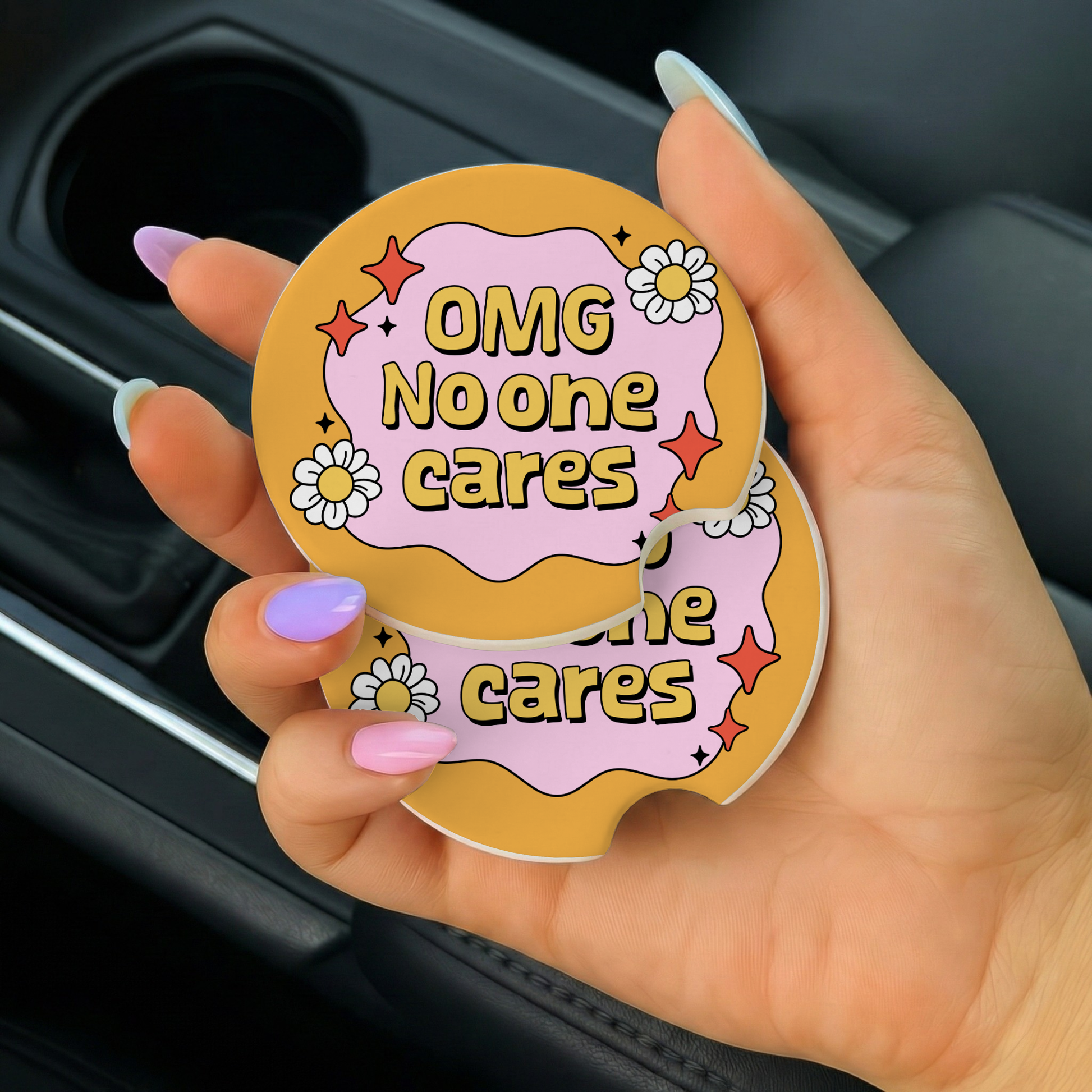 Hand holding a round, colorful coaster with text 'OMG No one cares' in a car.