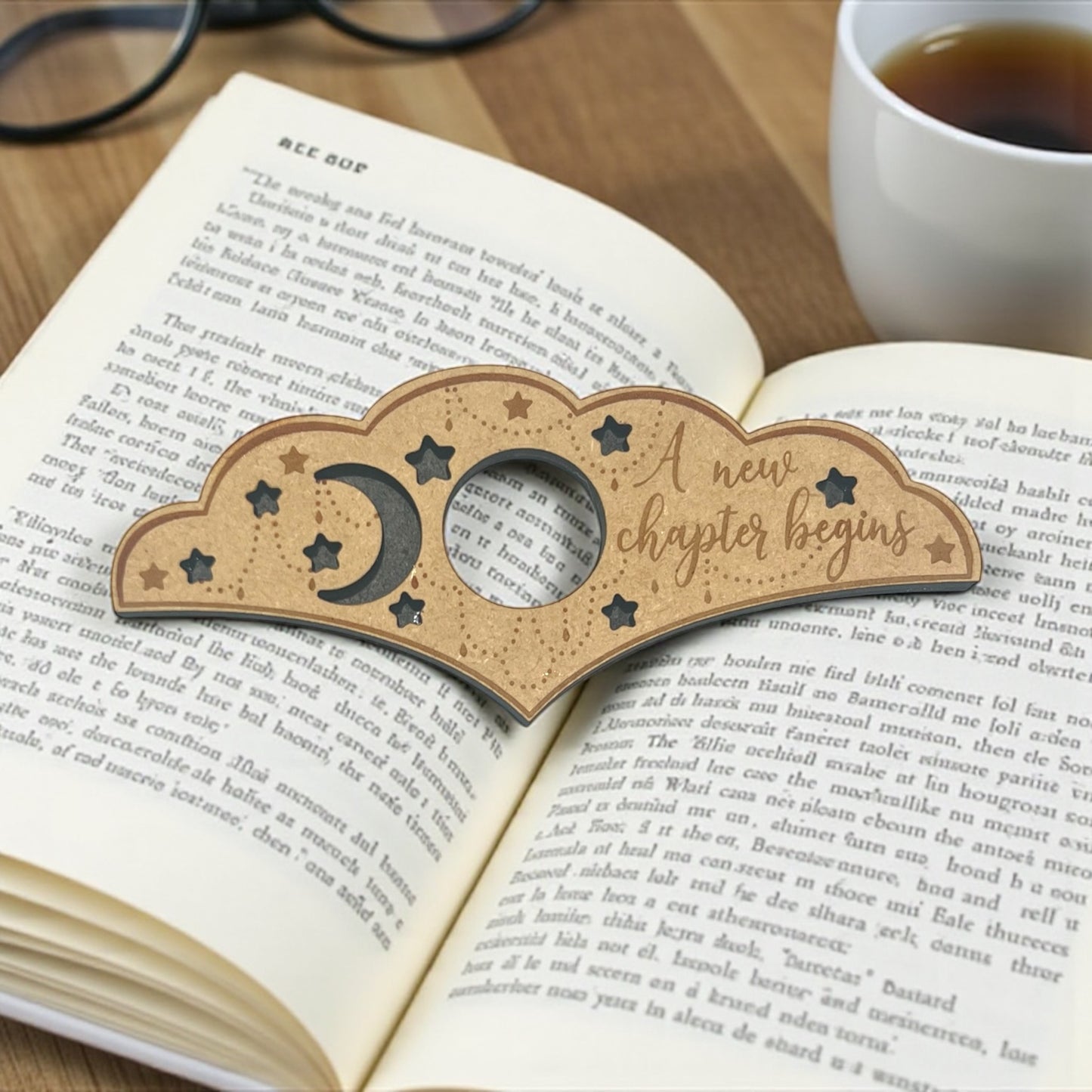 Wooden bookmark with 'A new chapter begins' on an open book next to a cup of tea.