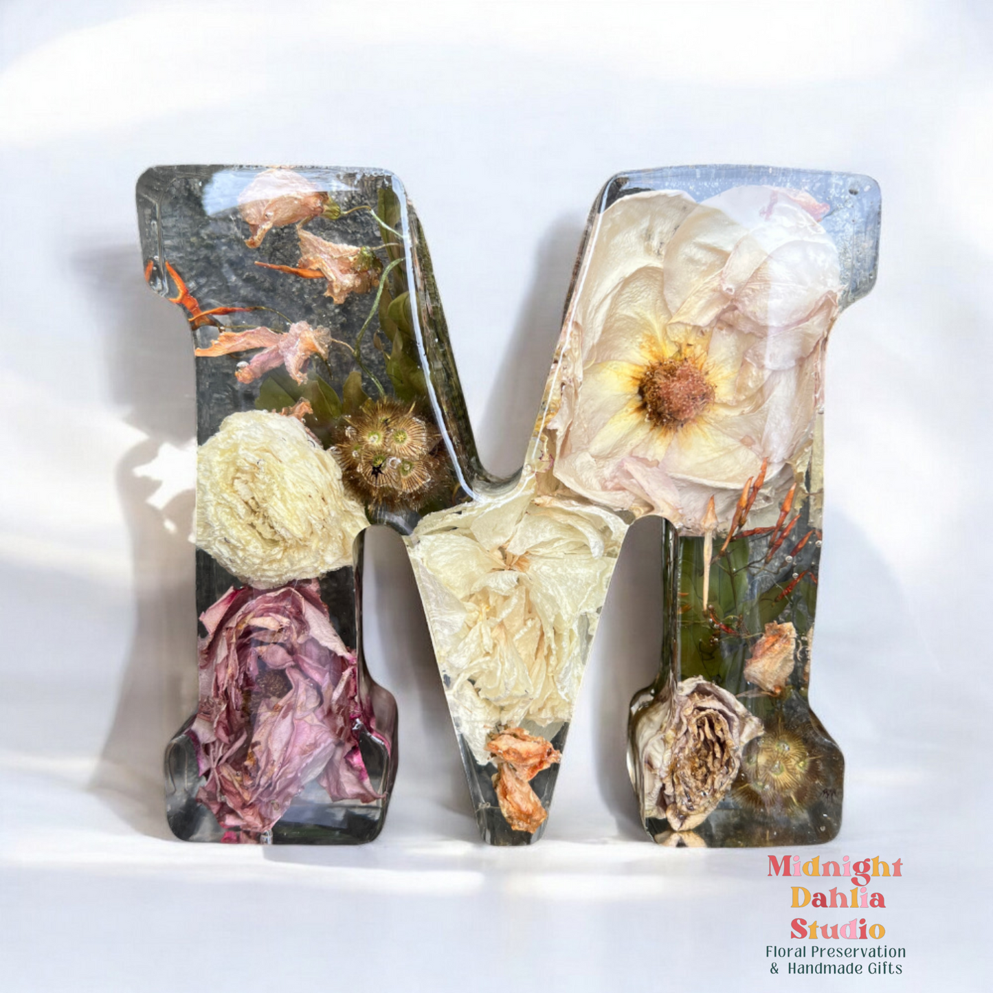 A crystal clear resin letter M with dried preserved flowers , including a rose , a ranunculus , kangaroo paw  , and green leaves.  A beautiful way to preserve your bridal bouquet.